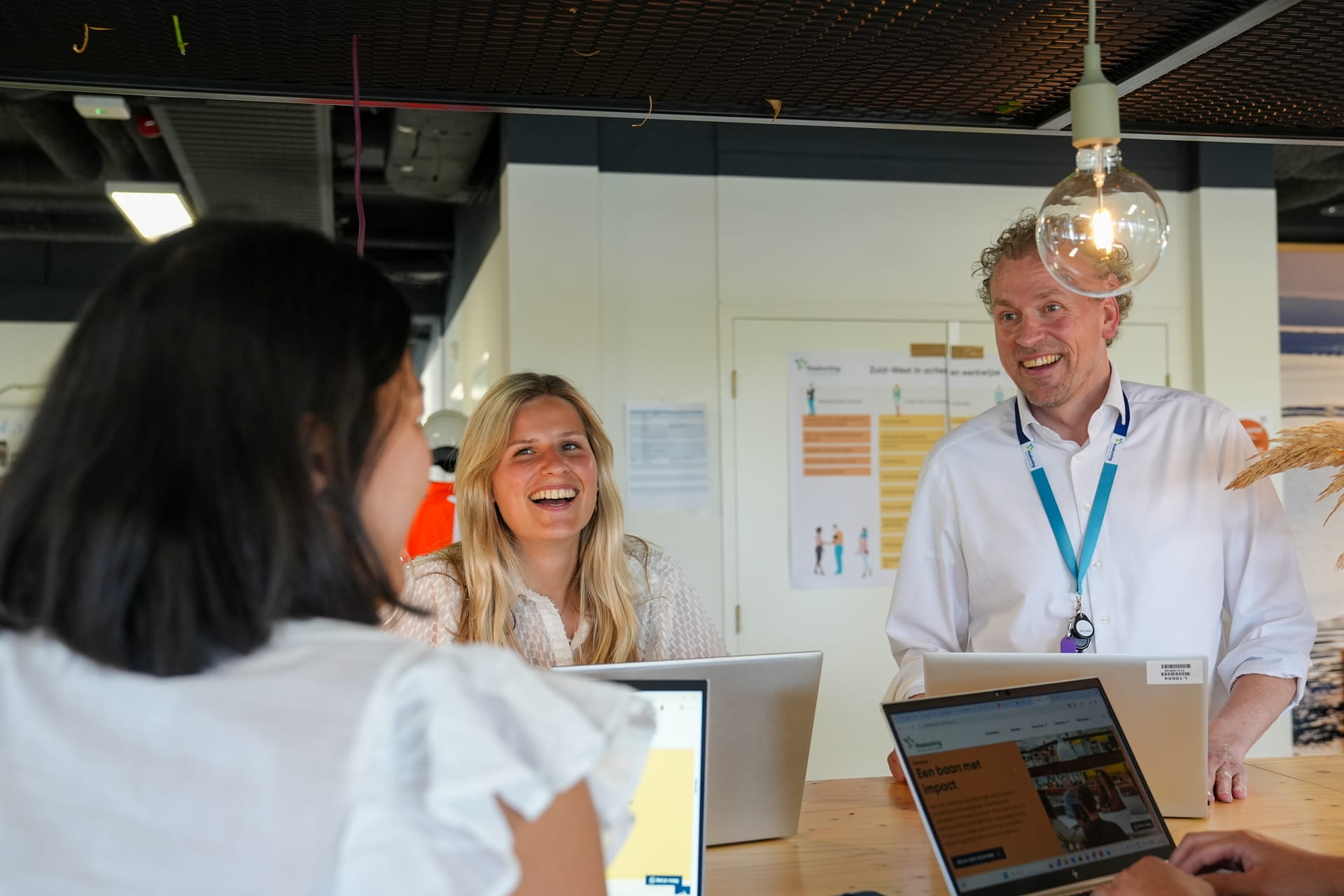 Drie lachende collega's in gesprek rond een hoge tafel met open laptops in een modern kantoor.
