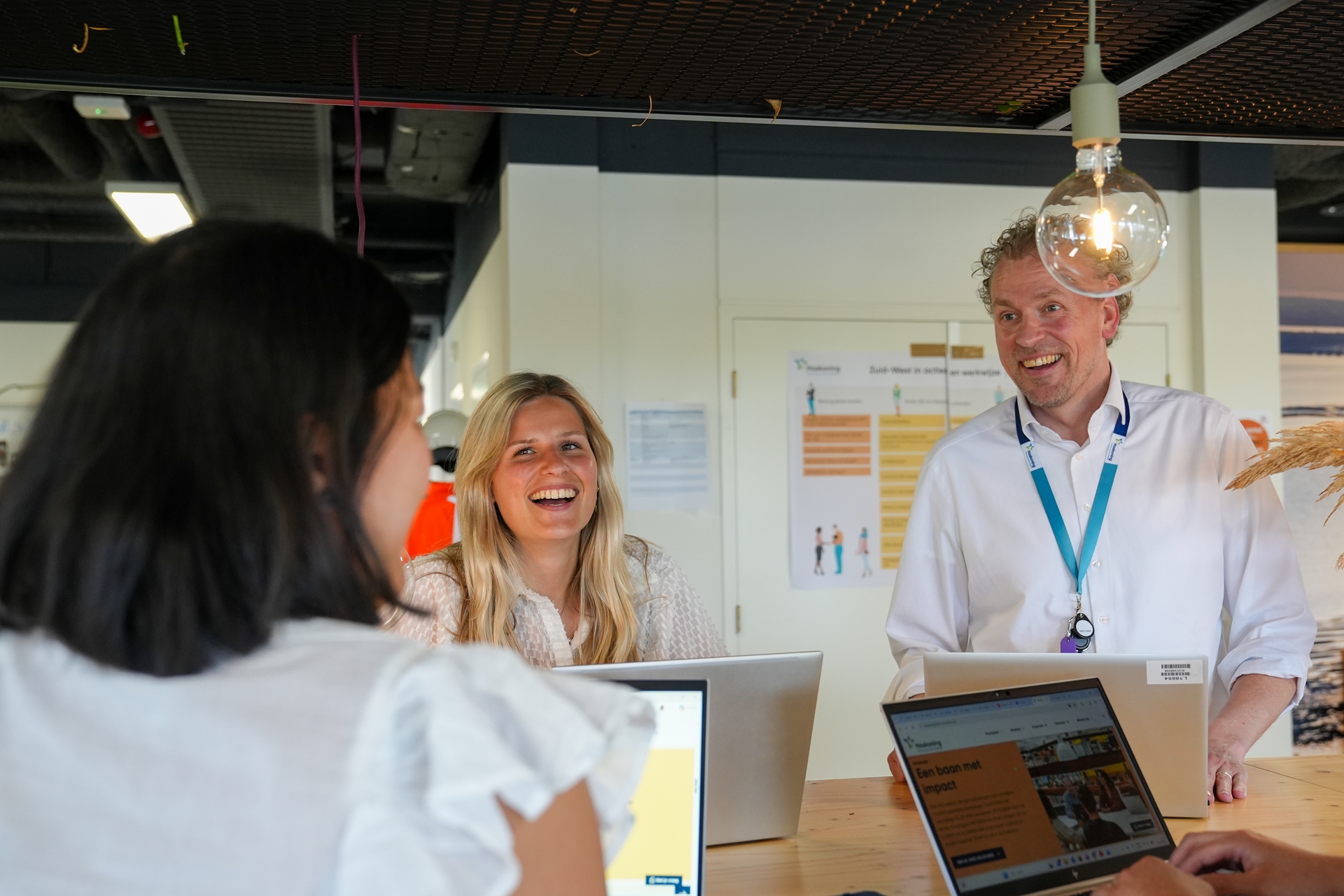 Drie lachende collega's in gesprek rond een hoge tafel met open laptops in een modern kantoor.