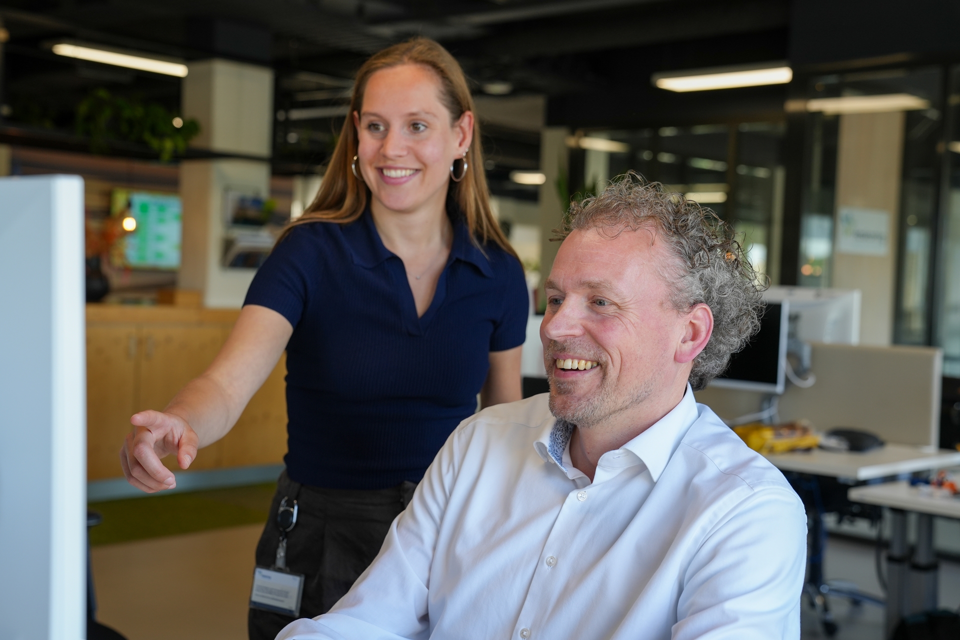Een vrouw die achter een mannelijke collega staat en glimlachend naar een beeldscherm wijst.