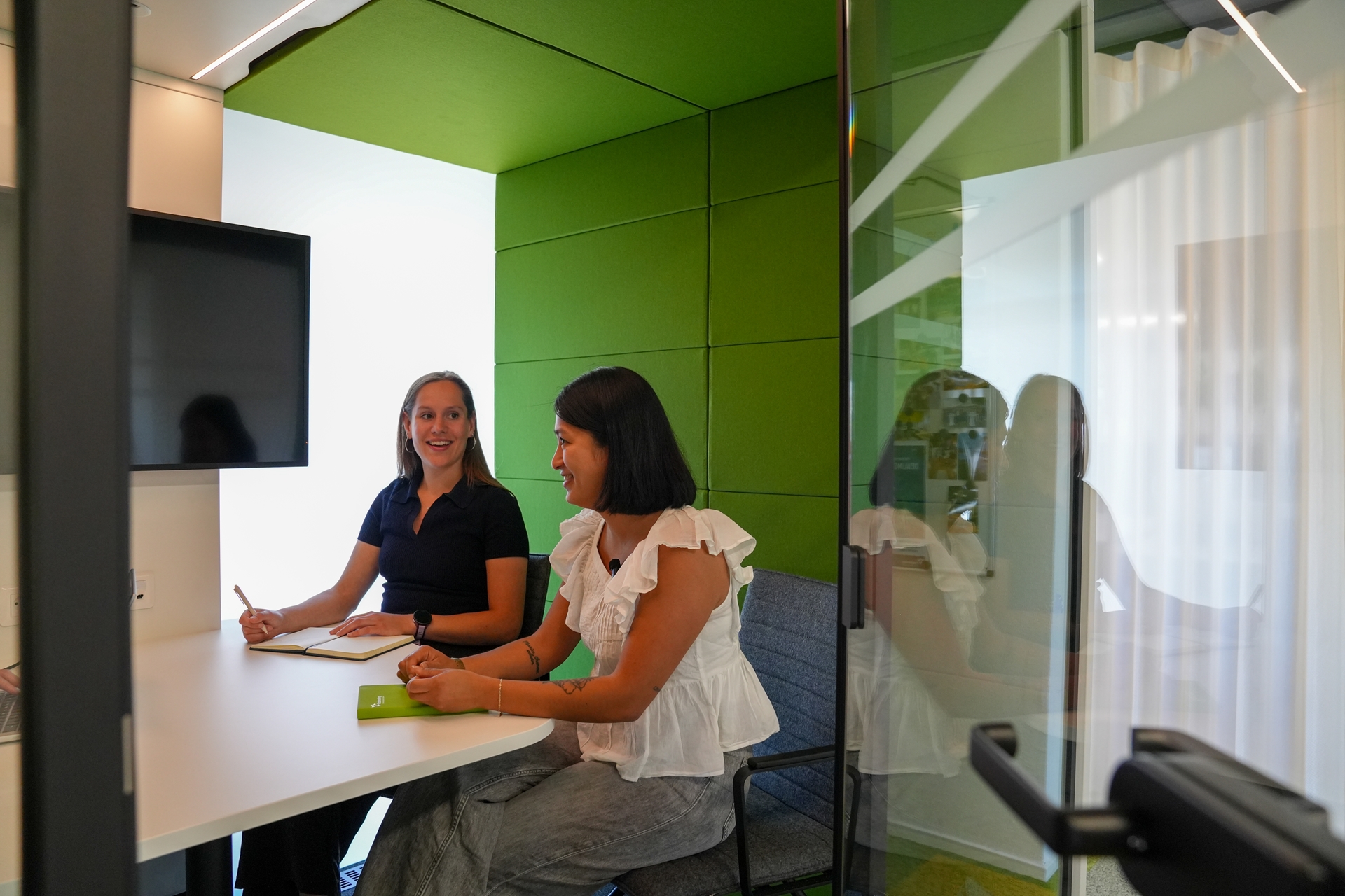 Twee vrouwen in gesprek aan een bureau in een groene overlegcabine met aantekenboeken.