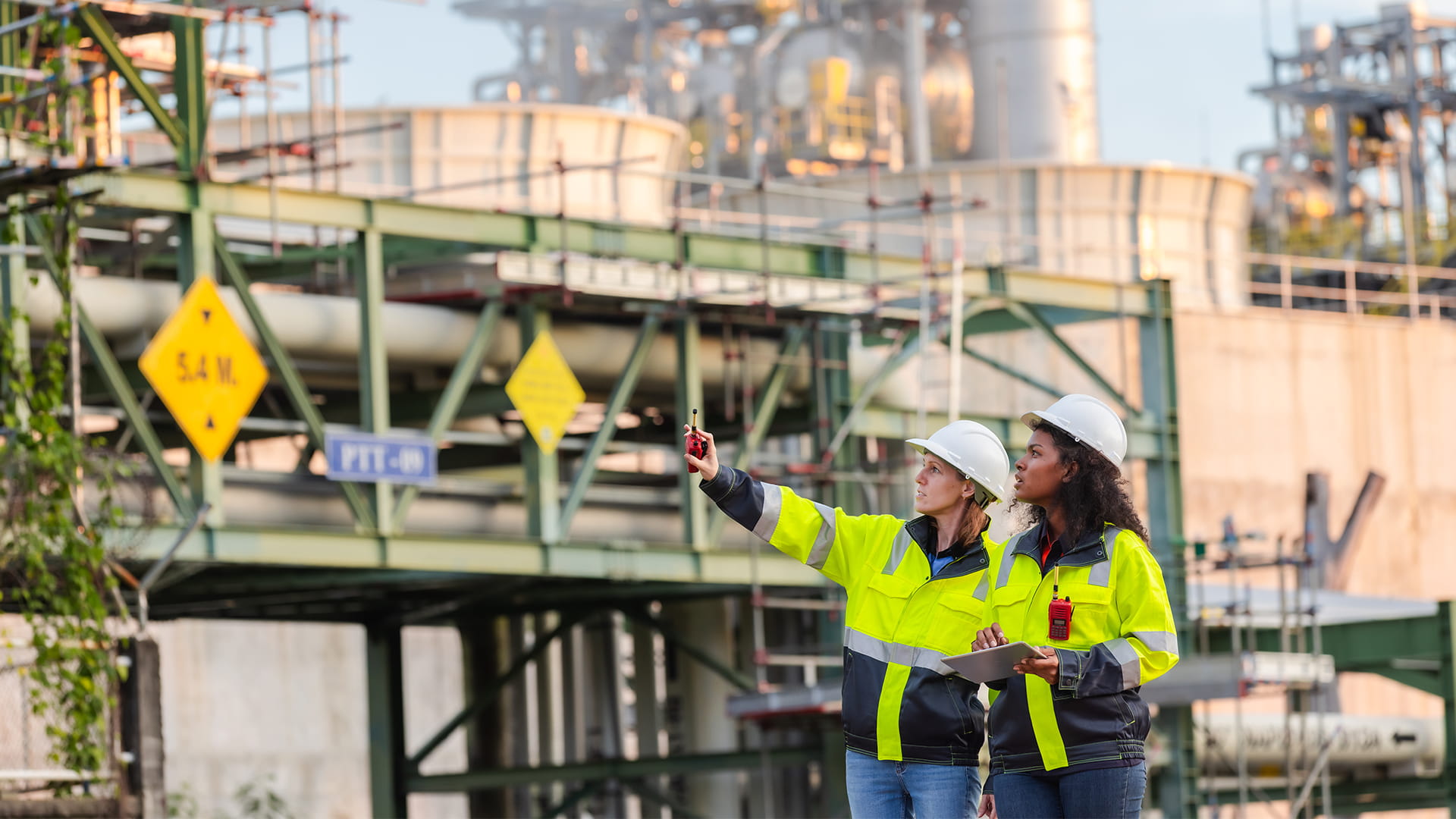 Twee professionals in veiligheidskleding en helm inspecteren een industriële installatie en wijzen onderdelen van de infrastructuur aan.