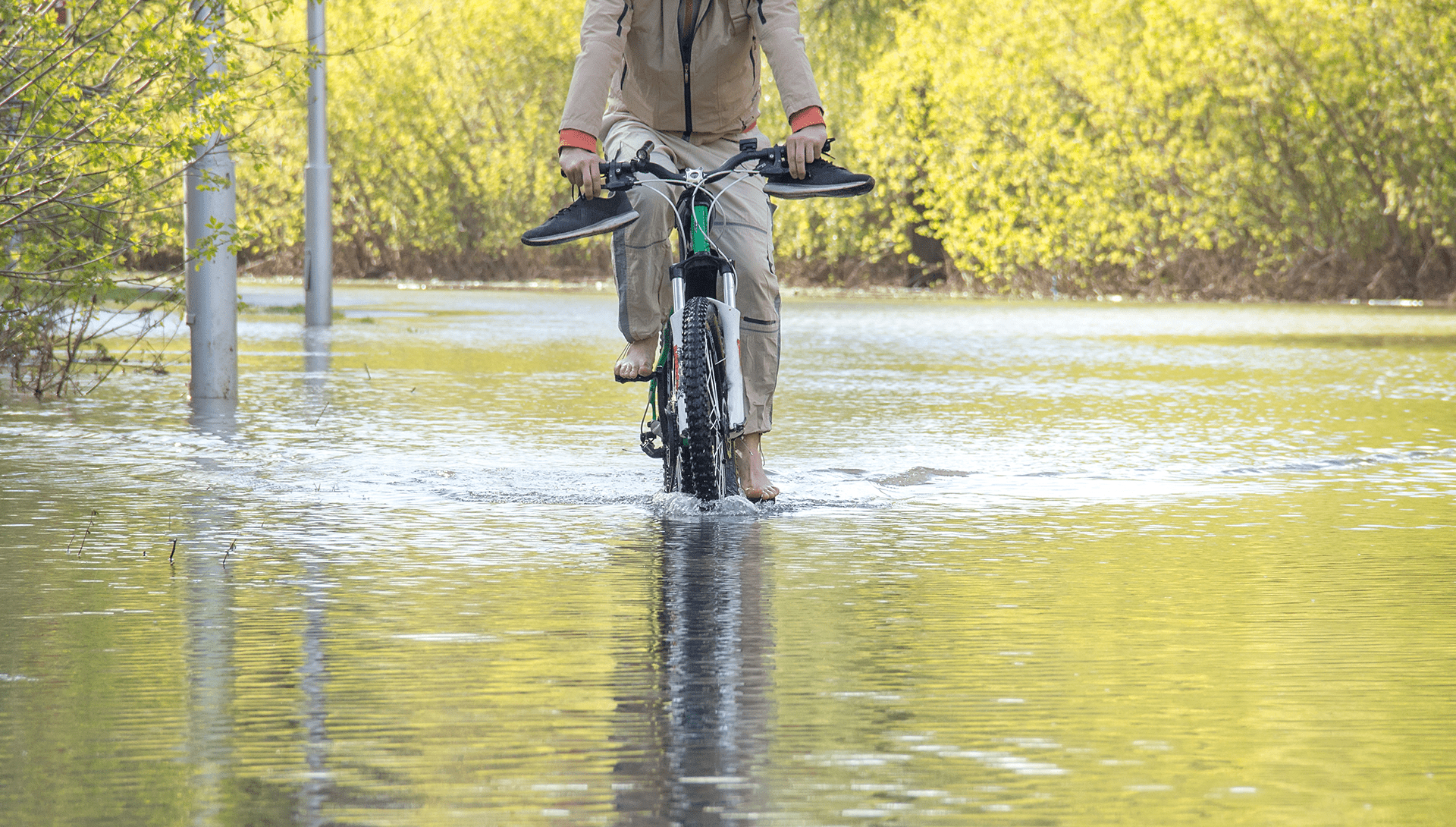 Fietser rijdt door water in een ondergelopen straat met schoenen in de hand