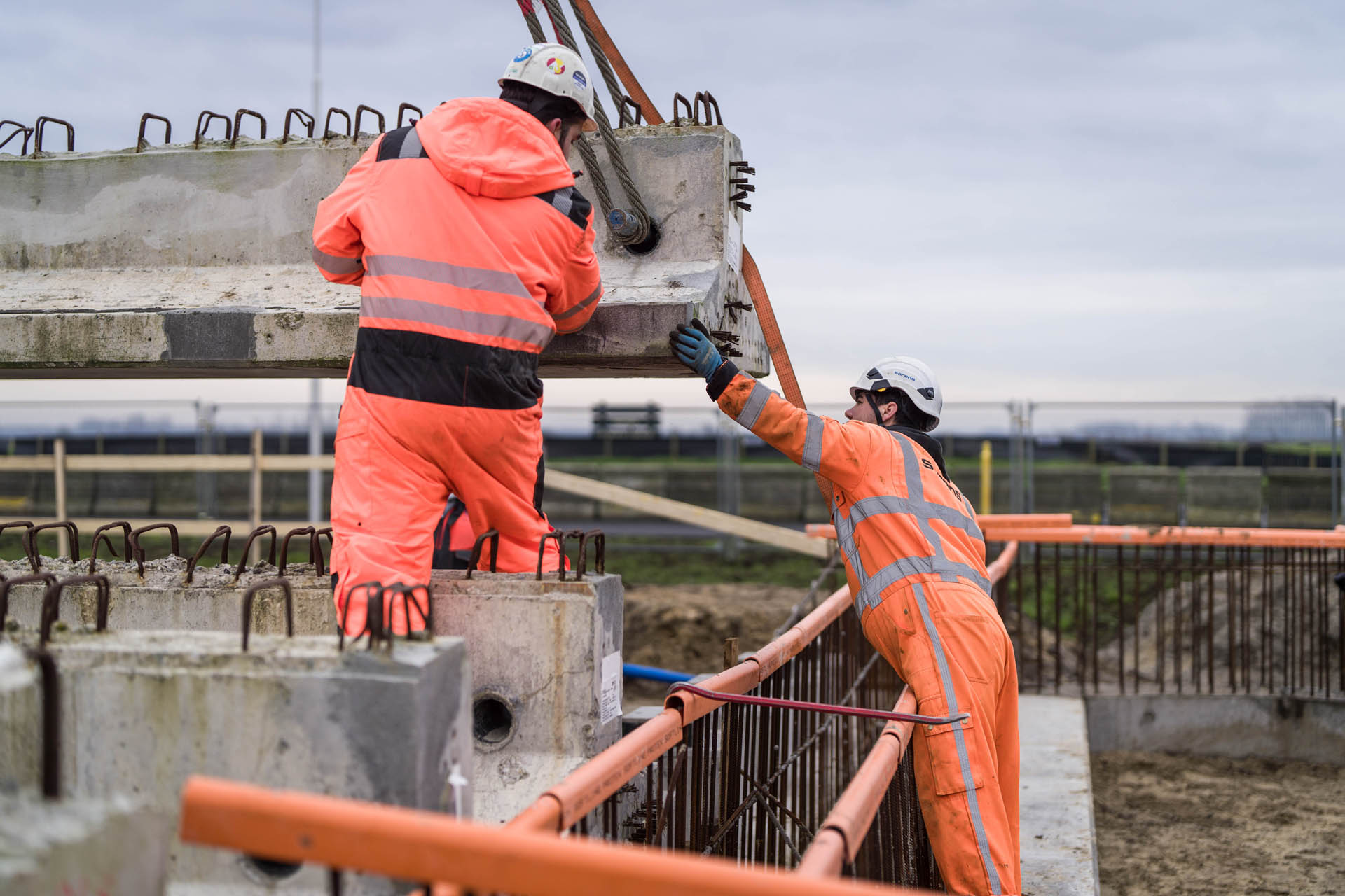 Twee bouwvakkers in oranje veiligheidskleding begeleiden een grote betonnen balk die door een kraan wordt opgetild.
