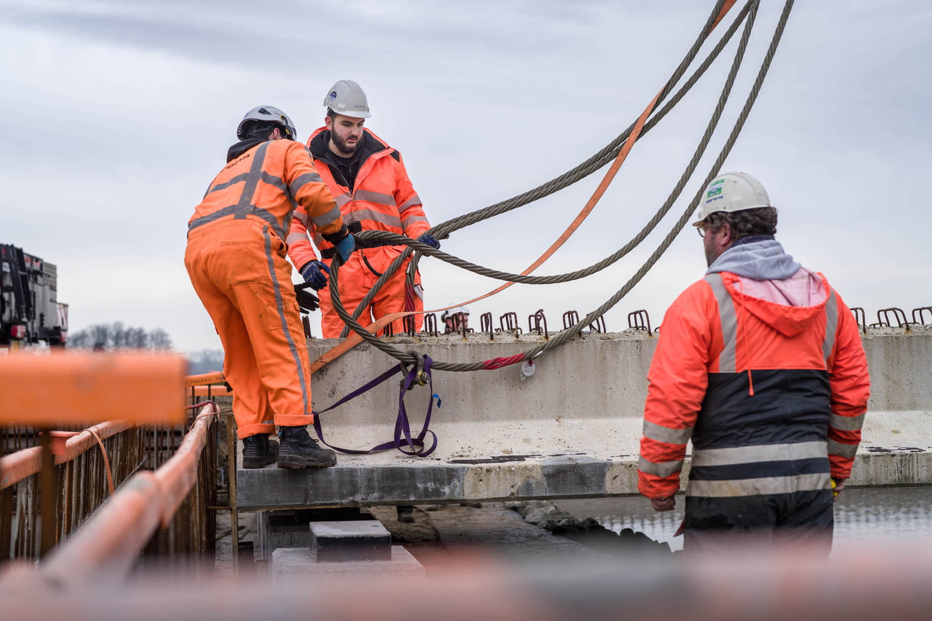 Drie bouwvakkers in feloranje jassen hanteren hijskabels die zijn bevestigd aan een grote betonnen balk op een bouwplaats.