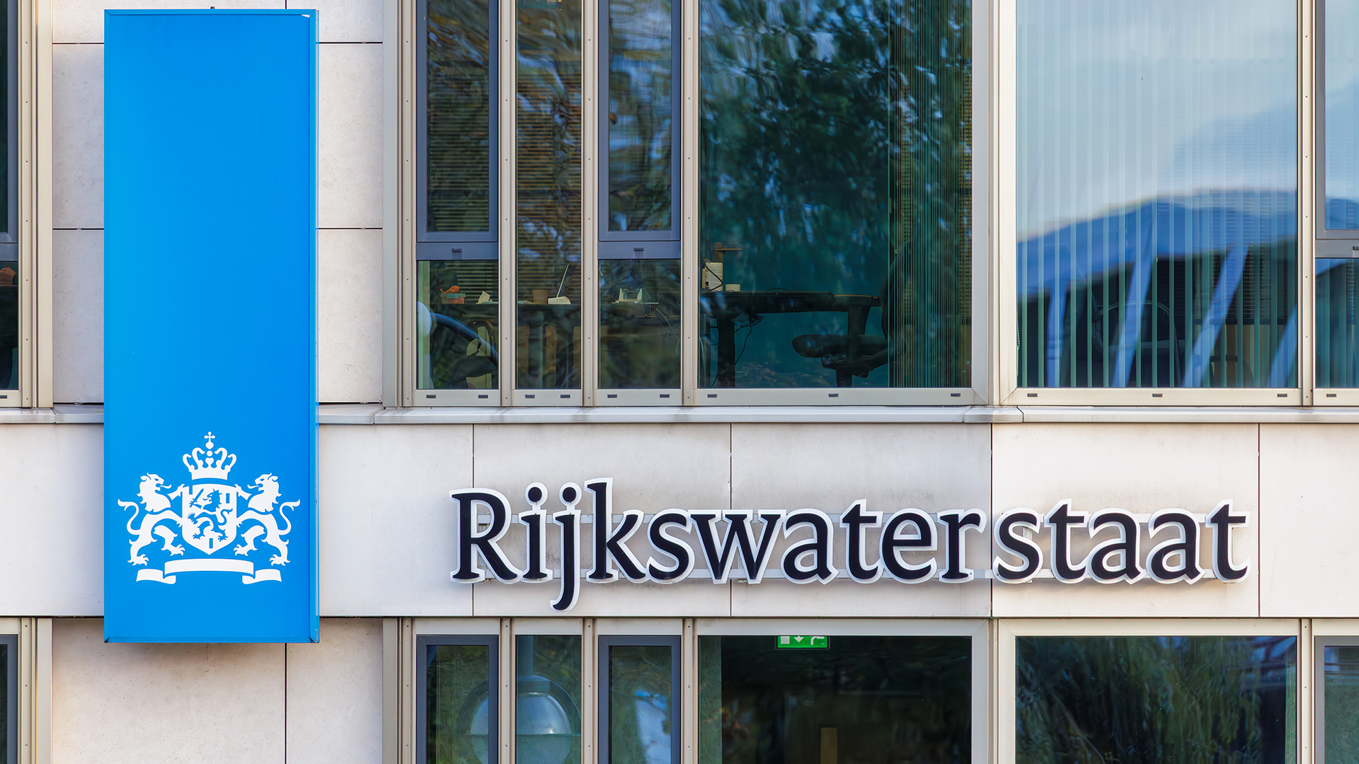 Gevel van een kantoorgebouw met een blauwe banner met het Nederlandse rijkslogo en de naam ‘Rijkswaterstaat’ in grote letters op de muur.