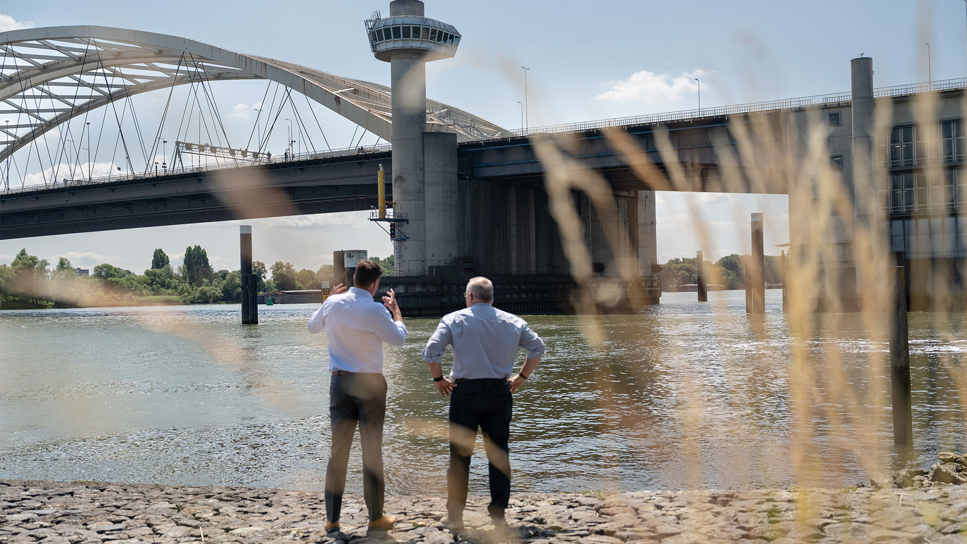Twee mensen die op de oever van een rivier staan en uitkijken richting de Van Brienenoordbrug in Rotterdam.