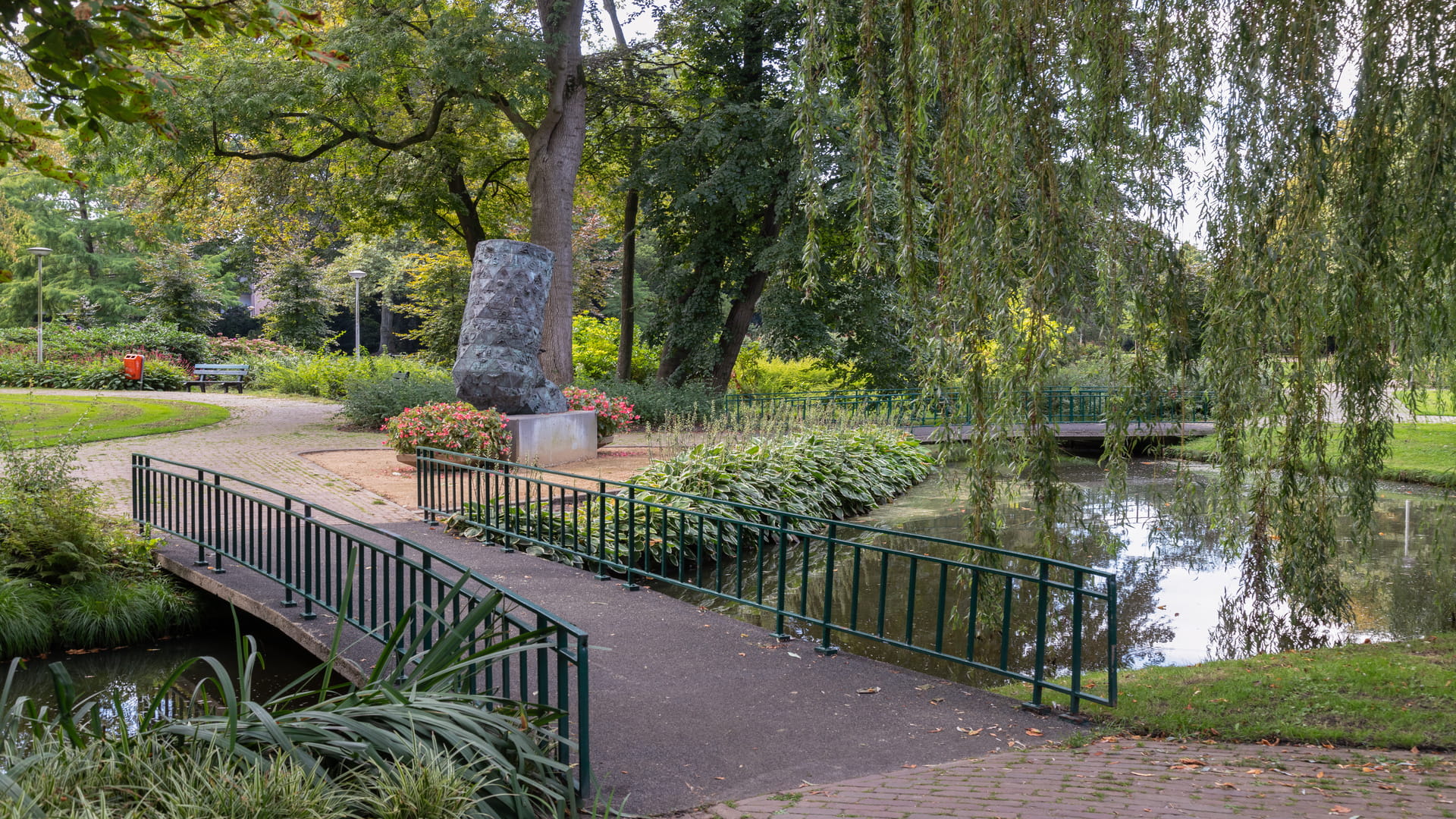 Voetgangersbrug over een smalle vijver in een groen park, omringd door bomen, struiken en een wandelpad met een kunstwerk op de achtergrond.