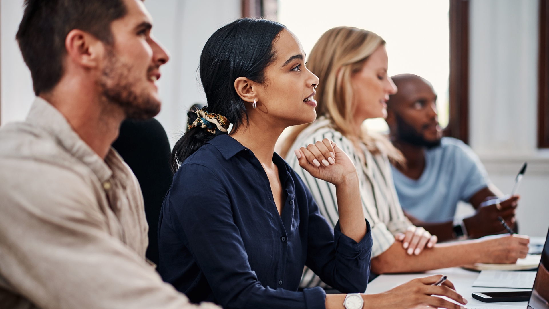 Een groep mensen zit aan een tafel tijdens een training. Ze luisteren aandachtig, maken aantekeningen en werken met een laptop.